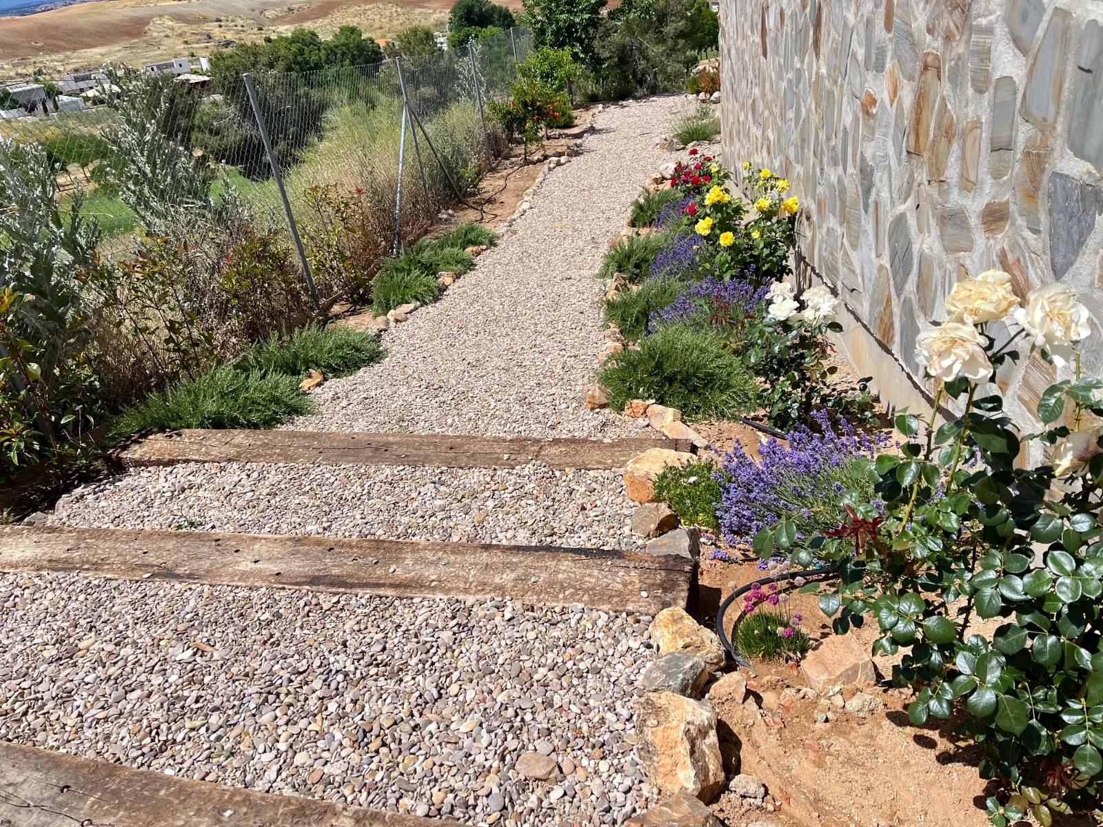 Sendero de grava con traviesas de madera y flores junto a un muro de piedra